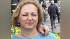 Brigitta Rasuli is smiling as she stands near a pavement and a public garden with daffodils. She has short, light brown hair and wears earrings and a blue top. The zoomed-in photo shows someone's arm around her shoulder.