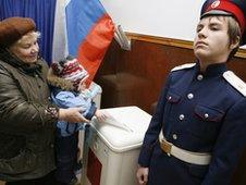 A woman carrying a child casts her ballot next to a Russian cossak (R) at a polling station in Rostov-on-Don