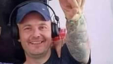 Close-up image of Wayne Neville, he's smiling with one arm pointing towards the camera. He's wearing a blue baseball cap with a pair of DJ headphones over the top. 