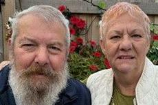 A couple poses together, a man with grey hair and beard, and a women with pink and grey hair. She has her arm around him.