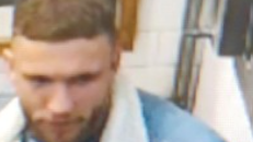 A CCTV image of a person wearing a blue denim jacket with a white, fluffy shearling-style lining along the collar and inner edges. Underneath the jacket, there is a teal or blue fitted top. A black necklace with a cross pendant is visible around the neck area. The person is also wearing dark pants and light-coloured sneakers with a patterned design.