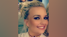 Cherry Turner with blonde hair in a ponytail, and makeup on, smiling at the camera, wearing a gold choker and a white top. 
