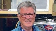 A picture of Dennis Chick. He is standing in front of a vehicle. He is smiling and wearing glasses. 