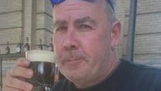 A close-up of a middle-aged man who is holding an Irish coffee in a glass. He is wearing a dark top.