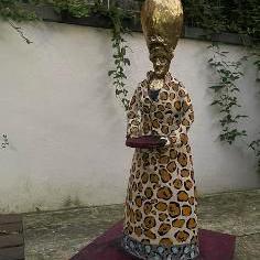 A gold-coloured statue of the goddess Ninkasi, on a purple coloured plinth. The statue is stood in a courtyard, with a white-painted wall behind which has some vegetation hanging down it.