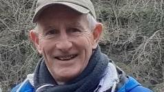 Mike Hutchinson wearing a hat, scarf and coat stood against a grey coloured bush