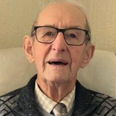 The head and shoulders of an elderly man wearing dark-framed glasses, a black and grey patterned cardigan, grey jumper, shirt and tie. He is sitting on a cream sofa and there is cream wallpaper behind him.