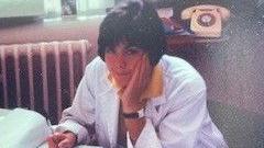 In this photograph from the 80s, Dr Olwen Williams is pictured at a desk. She's wearing a white overcoat with a yellow shirt underneath. She has a short black bob with a fringe.
