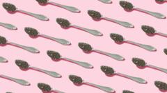 Diagonal silver teaspoons with chia seeds on them on a pink background.