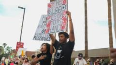 Fotografia colorida mostra um homem e uma mulher negros protestando com cartazes e diversas pessoas brancas protestando em segundo plano