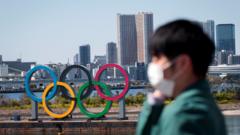 Tokyo Olympic rings in the background with a person wearing a face covering in the foreground