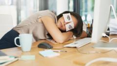 woman asleep at work, with head resting on her desk and post it notes on her eyes pretending to be awake
