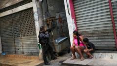 Sentados em frente a loja fechada, mãe e filho observam policial ao lado apontando arma grande