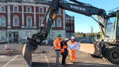 three people in construction clothing standing beneath a digger in a car park in front of a historic building