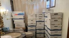 Boxes of papers are stacked in a bathroom with a chandelier and a toilet visible, at Mar-a-Lago