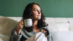 A woman looking into the distance while sat on a bed holding a cup