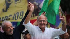 Fotografia colorida mostra Geraldo Alckmin, um homem branco idoso, meio careca, de óculos e nariz fino, segurando a mão de de Lula em frente a uma bandeira do Brasil