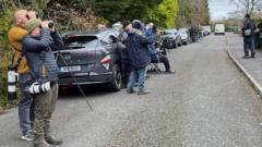 People and cars lined along the road, people have cameras and are looking through binoculars