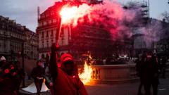 A demonstrator with a masked face holds a red flare in front of a pile of burning wood during protest in Paris
