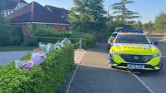 The scene at Carnoustie Drive in Great Denham. On the left is a hedge which has plastic-wrapped flowers sticking into it, and beyond is an entrance to a driveway with police tape across it. On the right are two police cars parked partly on the pavement. Beyond it is another detached house and trees.