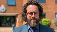 A detective inspector stands outside police HQ, he has long dark hair and a beard. He has red-rimmed glasses.