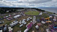 An aerial view of a highland games showing a parade ring, hundreds of people, and fairground rides.
