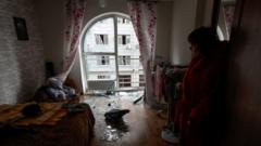 A woman stands in a room of her apartment with shattered window glass on the floor