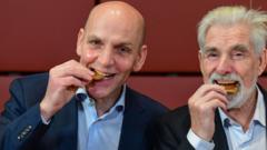 Laureate of the 2021 Nobel Prize in Chemistry, and Klaus Hasselmann, Laureate of the 2021 Nobel Prize in Physics, pretend to bite into chocolate versions of the gold medal awards as they attend the livestreamed Nobel Symposium at the Swedish Embassy