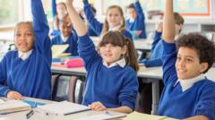 Children sit in rows at their desks in a classroom. All of them have their hands up. They are wearing white polo shirts and blue jumpers.