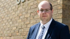 Jonathan Ash-Edwards standing outside a court brick building, looking at the camera. He wears thin-rimmed black glasses, a blue suit jacket, white shirt and blue and white striped tie. He is clean shaven with a balding head.