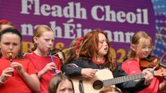 A group of young girls play instruments on a stage including a guitar, a fiddle, and tin whistles. They are standing on a stage in front of an Irish language sign which says Fleadh Cheoil na hÉireann.