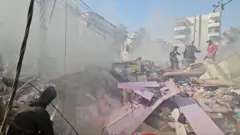 Recovery work in Dahieh, showing a large pile of debris, with a lot of pink crushed-up furniture. There's a lot of dust and emergency workers are standing on top of all the rubble.