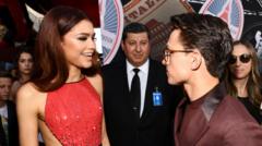 Zendaya and Tom Holland promoting Spider-Man: Far From Home together in 2019. She is wearing a red dress and he has on a purple suit as people look on in the background