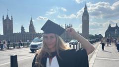 Melys stands in front of a London skyline with Big Ben and the Houses of Parliament behind her. She wears a cap and gown, with her hand holding the tip of her graduation cap.