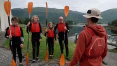 A dad and his three children - a girl and two older boys, stand in a line in water safety gear as an instructor gives a safety talk at the side of the loch. They are all holding bright orange paddles 