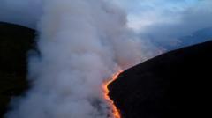 A drone image showing a bright zig-zag line of fire on a hillside at night. Thick smoke rises from the flames.