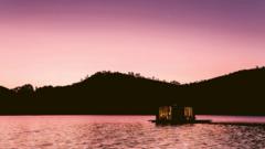Fotografia colorida mostra uma pequena casa em um lago com montanhas ao fundo durante um por-do-sol cor-de-rosa