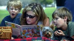 Deitados em gramado, mãe e dois filhos pequenos olham em direção a livro infantil