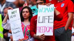 Two people hold up signs at the match - one reads "women, life, freedom"