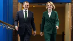 Katie Lam MP walks alongside Robert Jenrick. They are walking down a long corridor. Jenrick is wearing a dark suit, while Lam is wearing a brown suit. 