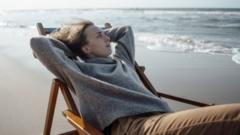Mulher deitada em cadeira em praia, com os braços atrás da cabeça e olhando para o "nada"