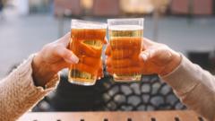 Close-up of two people toasting with pint glasses of beer while sitting at an outdoor wooden table.
