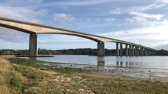 Orwell Bridge on a sunny, clear day. It is a long expanse of concrete with multiple columns that come down into the water. A boat is parked in the mud underneath one of the large arches. In the foreground is a sandy area with some grass.