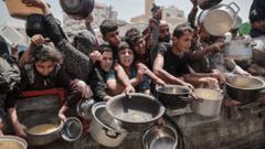 A group of children reach over a barrier with various pots and pans, seeking food or water. The crowd includes men and women of different ages, with many arms extended in a chaotic scene. The cookware varies in size and type, including metal pots, plastic containers, and ladles. The background features the urban buildings and structures of Gaza City.