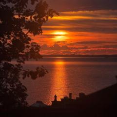 An orange sunrise with the bright beam reflected in the sea, rooftops and trees are silhouetted in the foreground.