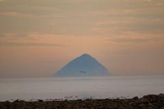 A cone-shaped island at sunset under a pastel-coloured sky, with three birds flying across the shot above the sea.