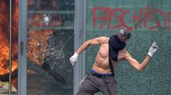 Manifestante sem camisa, de rosto coberto e boné, atira objeto em frente a uma fachada com vidros quebrados, refletindo um incêndio