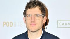 Elis, who has brown curly hair, stubble and wears clear plastic-frame glasses. He is wearing a white t shirt and navy shirt and is looking at the camera with a neutral expression.