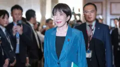 Sanae Takaichi in a blue blazer and black blouse, walking in a hallway