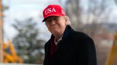 Donald Trump smiles to the camera while walking towards the White House from the main lawn. He's in a black coat and wearing a red cap with the writing USA in white at the front